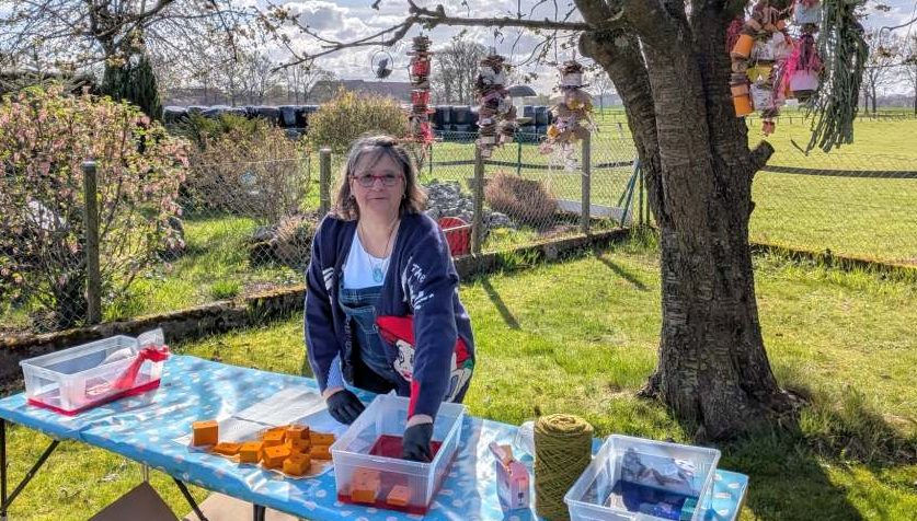 Susanne steht draußen und färbt mit lebensmittelechten Farben Balsaholzklötze. Im Baum hängt bereits fertiges Becherglück-Spielzeug.